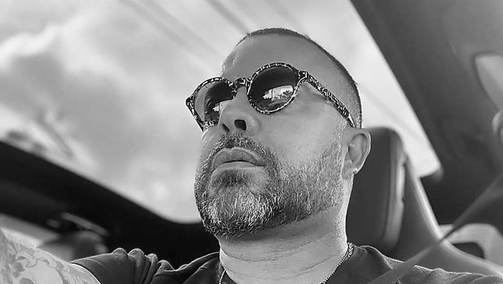 Ricardo Godoi sitzt in einem Auto und trägt eine Sonnenbrille mit auffälligem Design. Sein Gesichtsausdruck wirkt entspannt und konzentriert, während er geradeaus schaut. Das Bild ist in Schwarz-Weiß gehalten, was die Details seines Bartes und der Sonnenbrille besonders hervorhebt.