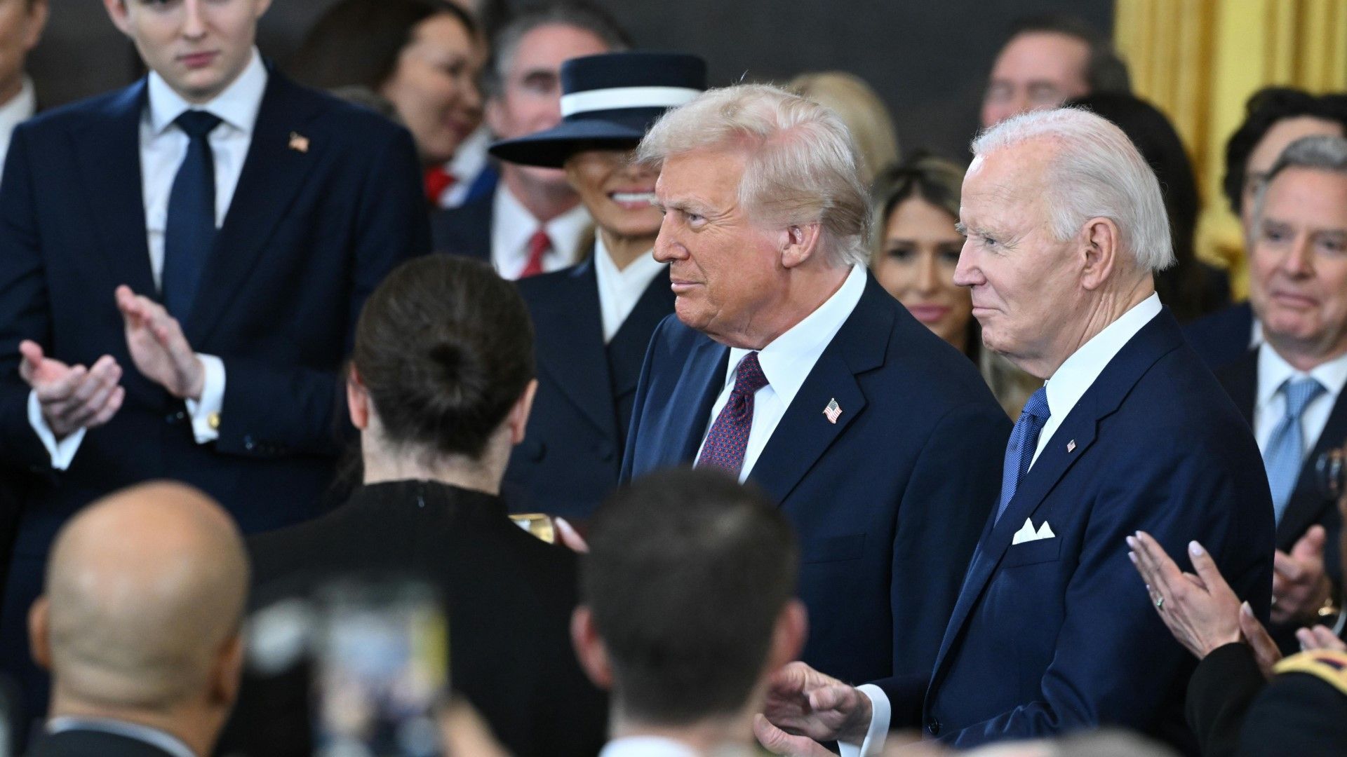 Donald Trump und Joe Biden stehen nebeneinander bei einer formellen Veranstaltung, umgeben von applaudierenden Gästen. Im Hintergrund sind mehrere Personen in eleganter Kleidung, darunter eine Frau mit Hut. Donald Trump und Joe Biden stehen nebeneinander bei einer formellen Veranstaltung, umgeben von applaudierenden Gästen. Im Hintergrund sind mehrere Personen in eleganter Kleidung, darunter eine Frau mit Hut.